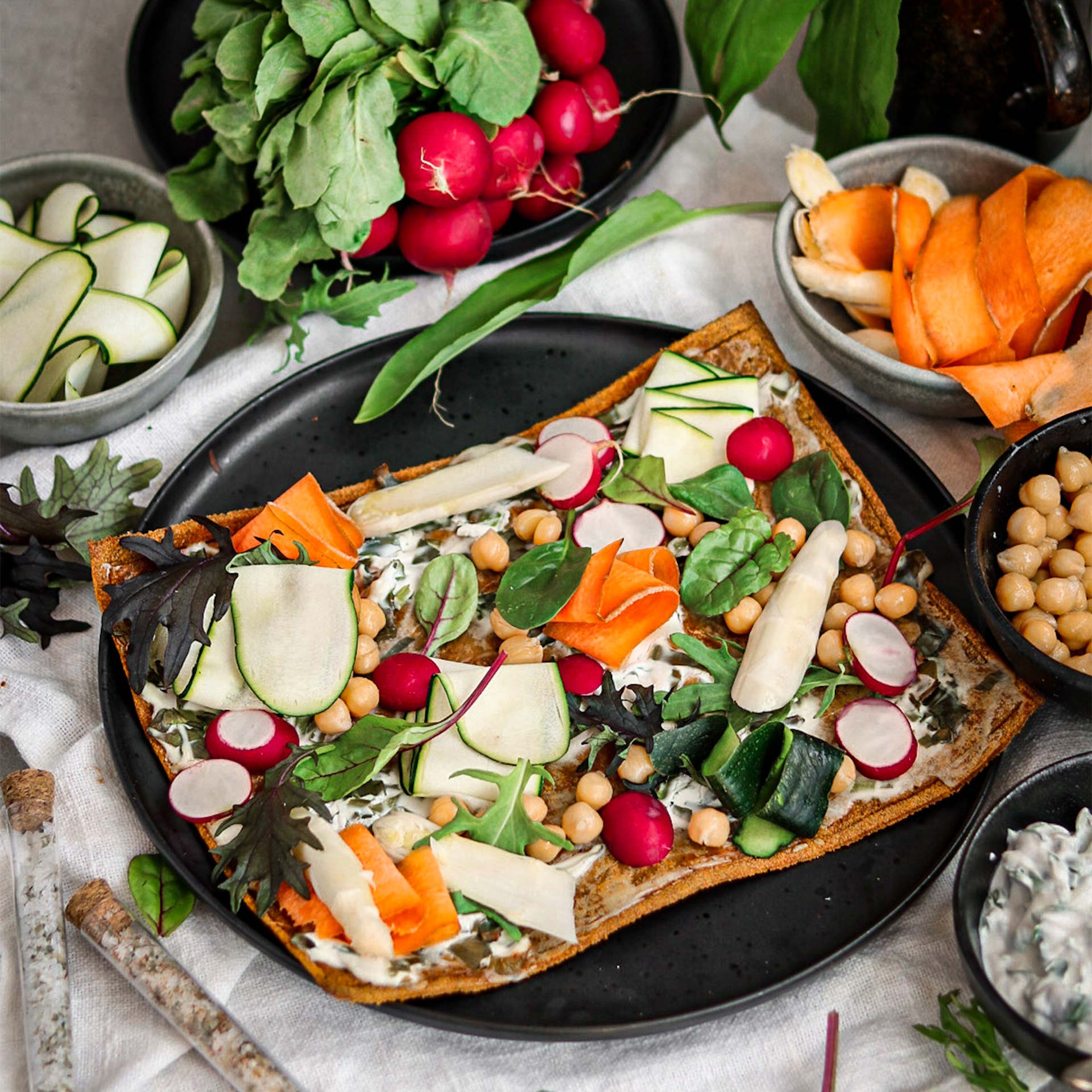 Gebackener Pizzaboden auf schwarzem Teller belegt mit Kichererbsen, Spargel, Zucchini, Radieschen, Karotten, und Salat